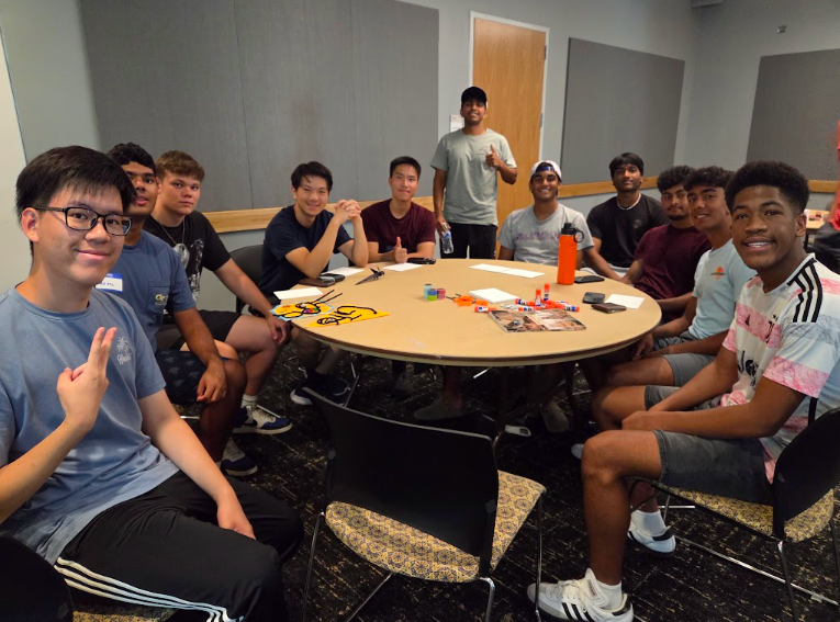 A group of approximately 11 students sits and stands around a round table in a classroom or meeting room. Craft supplies, papers, and personal items are spread across the table, and acoustic wall panels and a door are visible in the background.